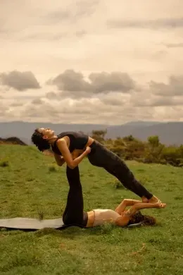 Couple practicing acroyoga together on a grassy mat, with cloudy skies and hills surrounding them near Unika Hotels