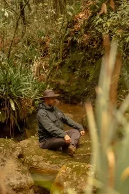 Man in a hat meditating cross-legged on a mossy rock, surrounded by ferns and a shallow forest stream near Unika Hotels