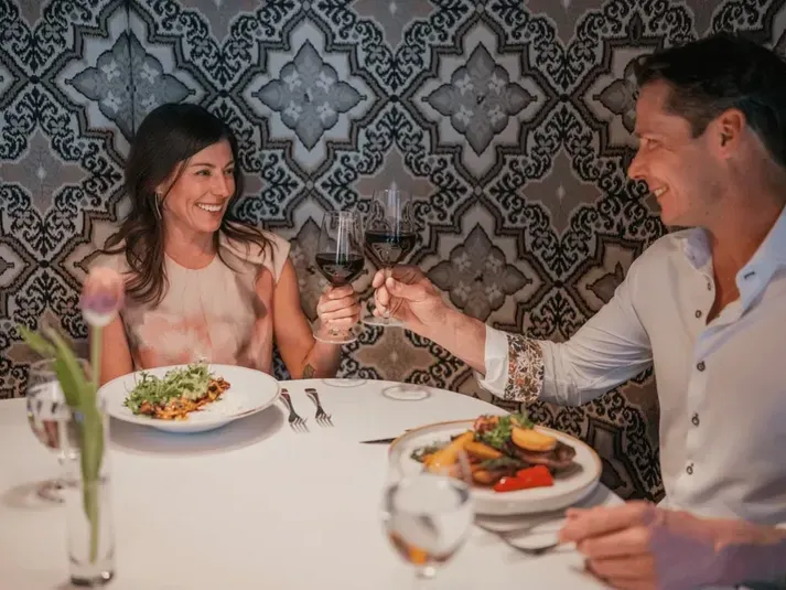 Couple at a restaurant, toasting with wine glasses, surrounded by plates of food and a decorative wall.