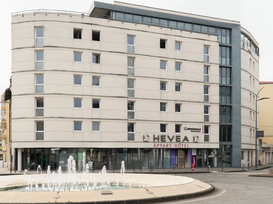 Exterior of Appart’Hôtel Hévéa with many windows and a fountain in front.
