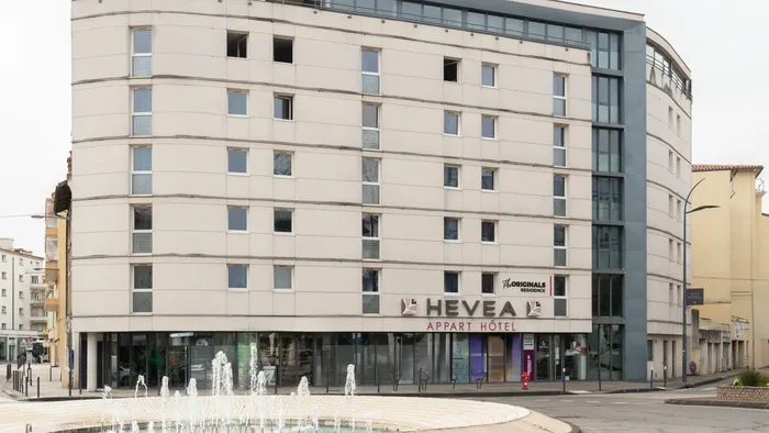 Exterior of Appart’Hôtel Hévéa with many windows and a fountain in front.