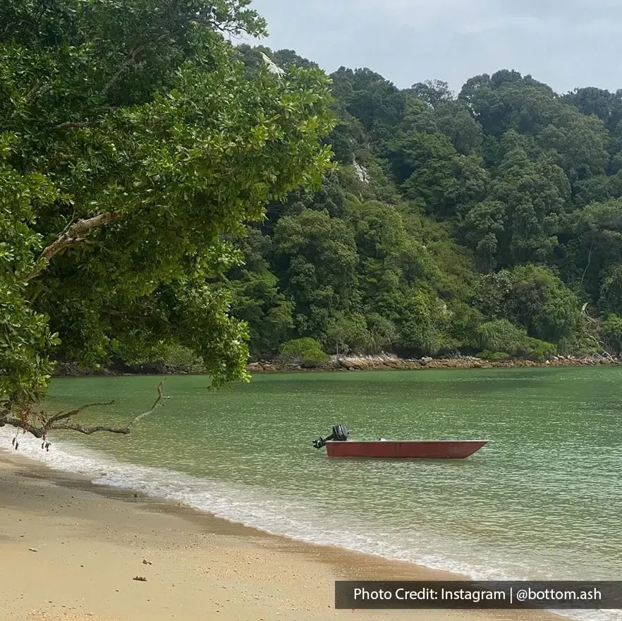 Pemandangan pantai tropika dengan pokok rendang di tepi laut dan bot merah di permukaan air - Lexis Hibiscus Port Dickson