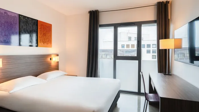 Room at Appart’Hôtel Hévéa with a bed, desk, chair, lamp, and large window.