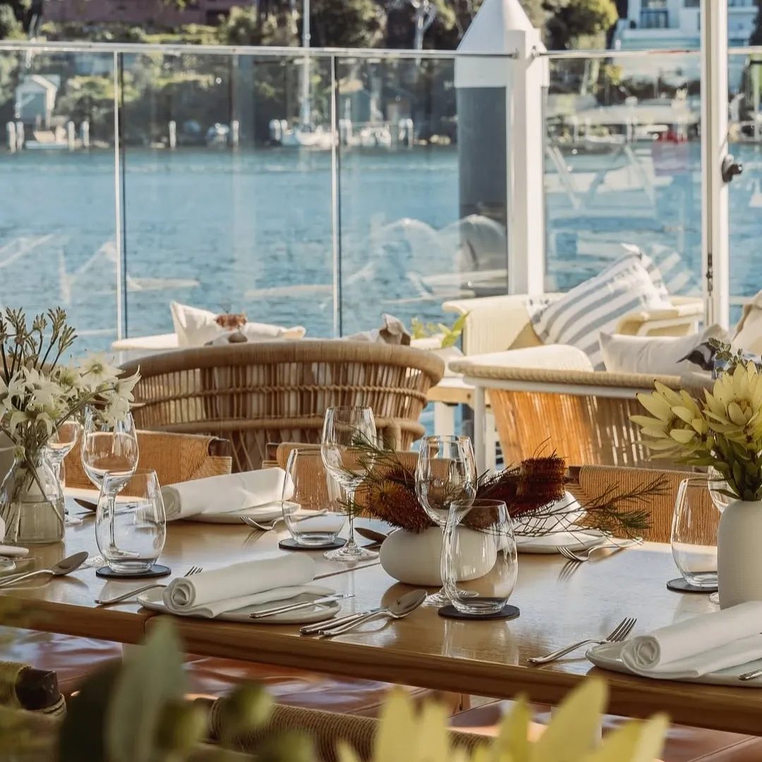 A table at Elysium Hotels is beside a glass railing overlooking the water with rattan chairs and native flora vase