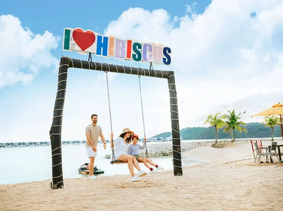 Family enjoying a swing on the beach under an I love Hibiscus sign.