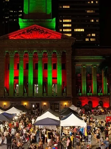 Festive night scene with illuminated Christmas tree and market stalls in front of a building with red and green lights