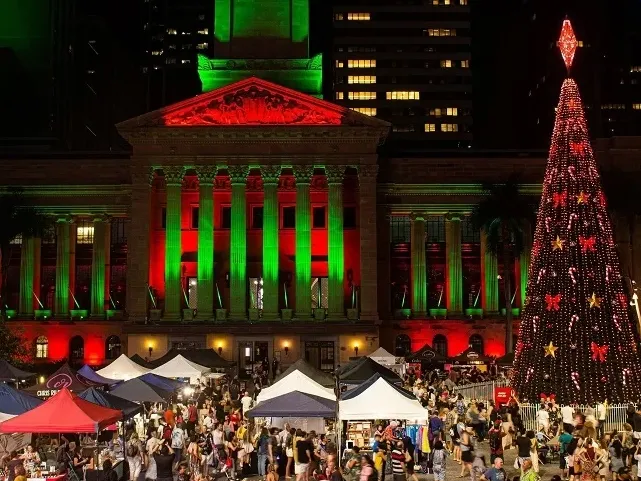 Festive night scene with illuminated Christmas tree and market stalls in front of a building with red and green lights