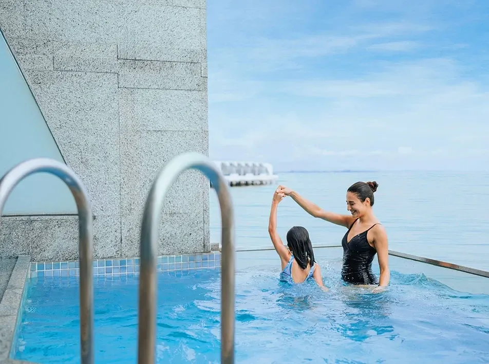 Mother and daughter enjoying a playful moment in a seaside infinity pool.