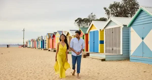 Couple walks past painted boathouses in Brighton Beach