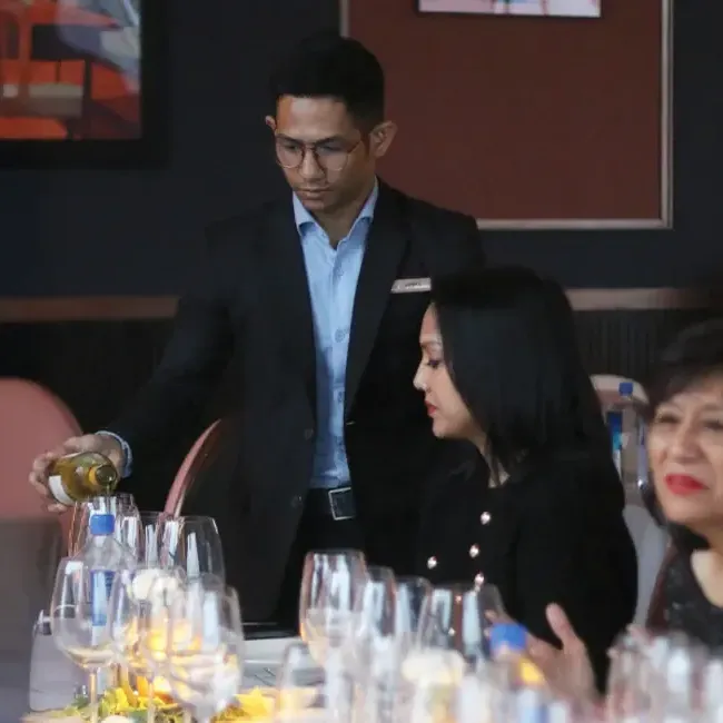 A man pours wine for guests seated at a dining table, creating a warm and inviting atmosphere - Lexis Hotels & Resorts