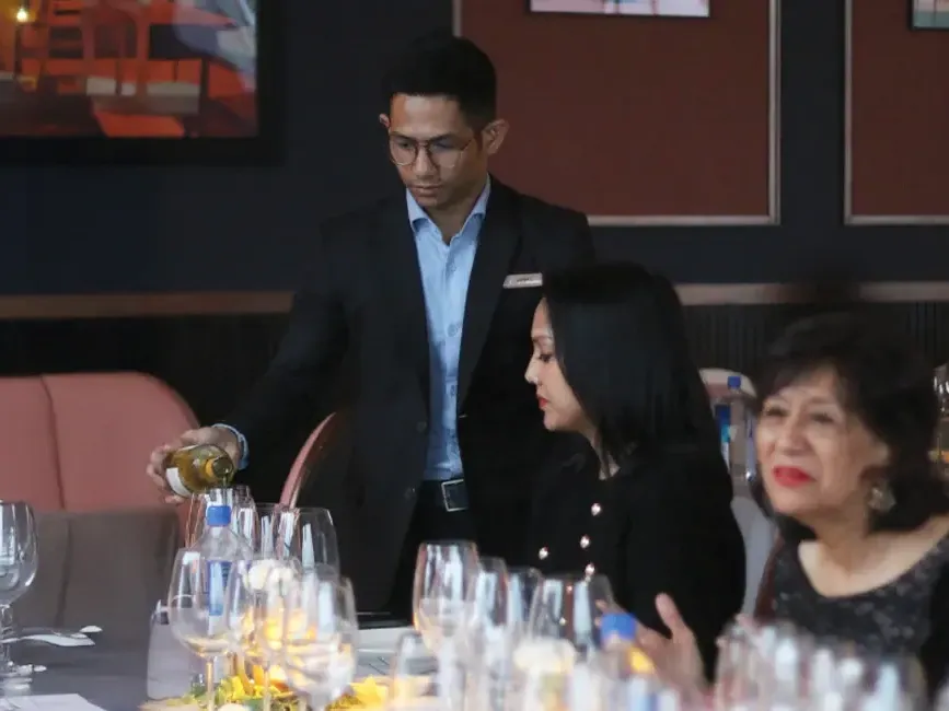 A man pours wine for guests seated at a dining table, creating a warm and inviting atmosphere - Lexis Hotels & Resorts