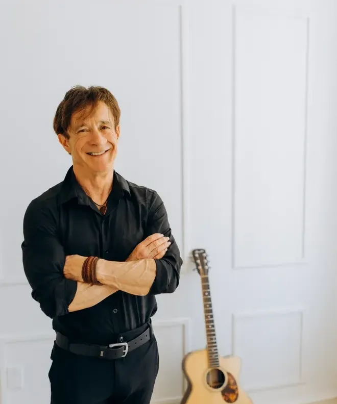 Dan Doiron standing with arms crossed next to an acoustic guitar against a white wall at Rodd Brudenell River Resort