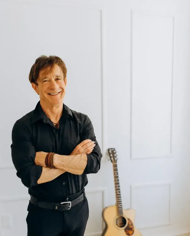 Dan Doiron standing with arms crossed next to an acoustic guitar against a white wall at Rodd Brudenell River Resort