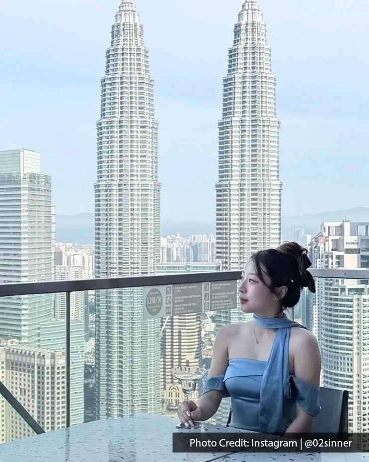 A woman enjoying a light drink at a bar overlooking Kuala Lumpur skyline, showcasing the Petronas Twin Towers.