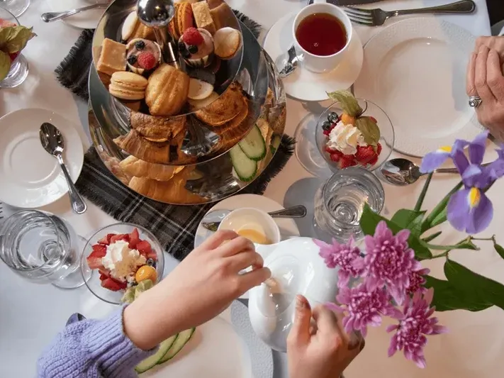 A table set with assorted pastries, flowers, and a person pouring tea into a teapot.