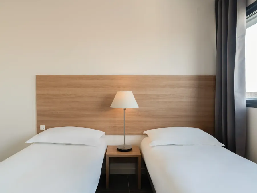 Two single beds with wooden headboard and nightstand lamp in a room at Appart’Hôtel Hévéa, Valence.