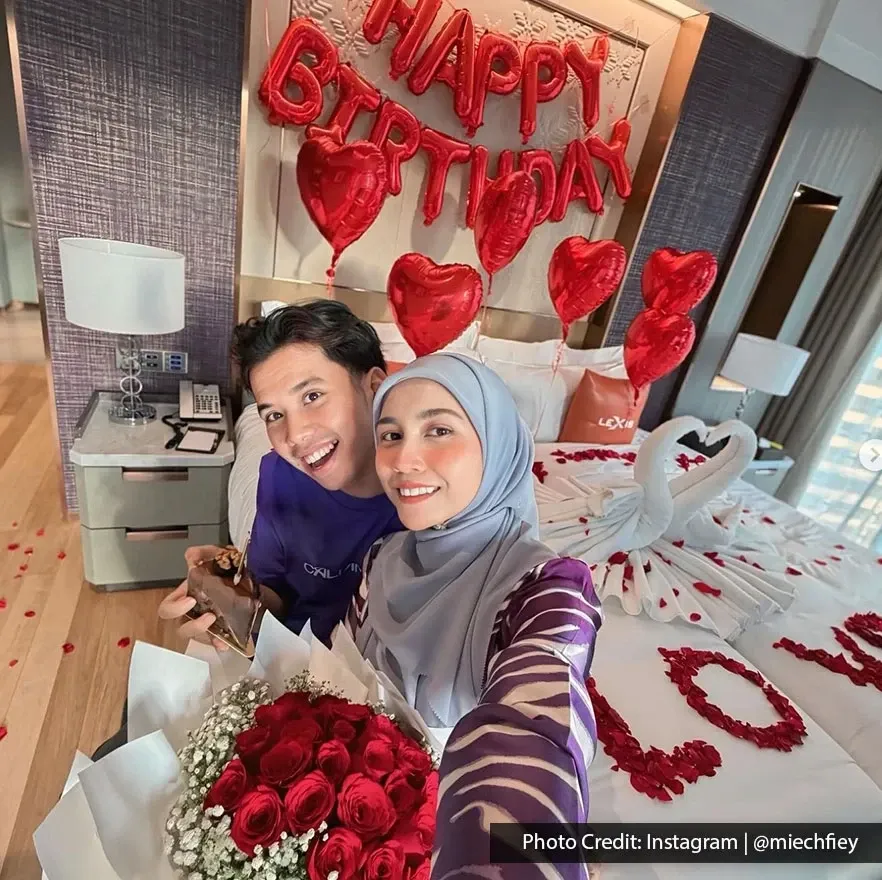 A couple smiles on a bed while taking a selfie in a decorated room with birthday balloons.