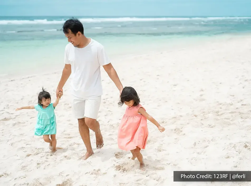 A father holding his daughters' hands while walking on the beach - Lexis MY