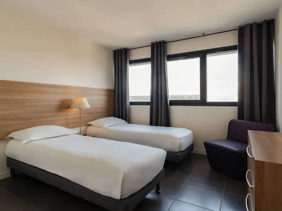 Two twin beds with headboards, a dresser, and a chair in a two-bedroom apartment at Appart’Hôtel Hévéa, Valence.