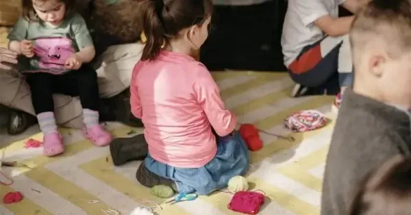 little girl in pink top kneeling on mat with knitting tools around her. children in foreground