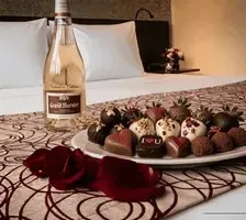Wine bottle beside chocolate-covered strawberries with red rose petals on a patterned bed runner at Unika Hotels