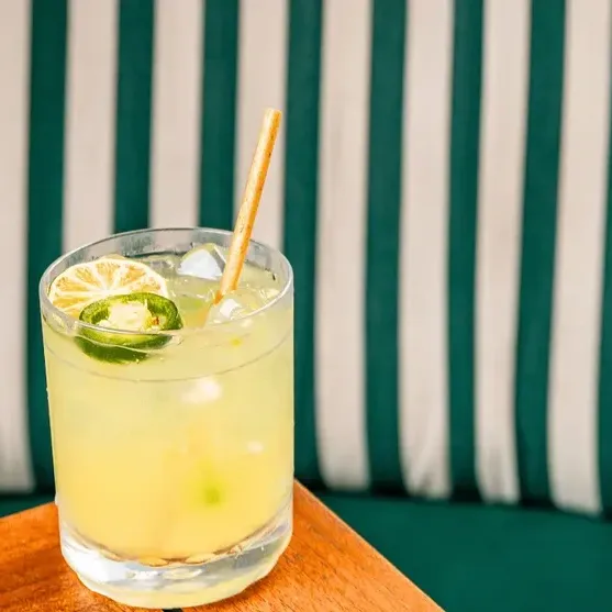 A glass of yellow cocktail with a straw and lime slices, placed on a table with a green striped background.