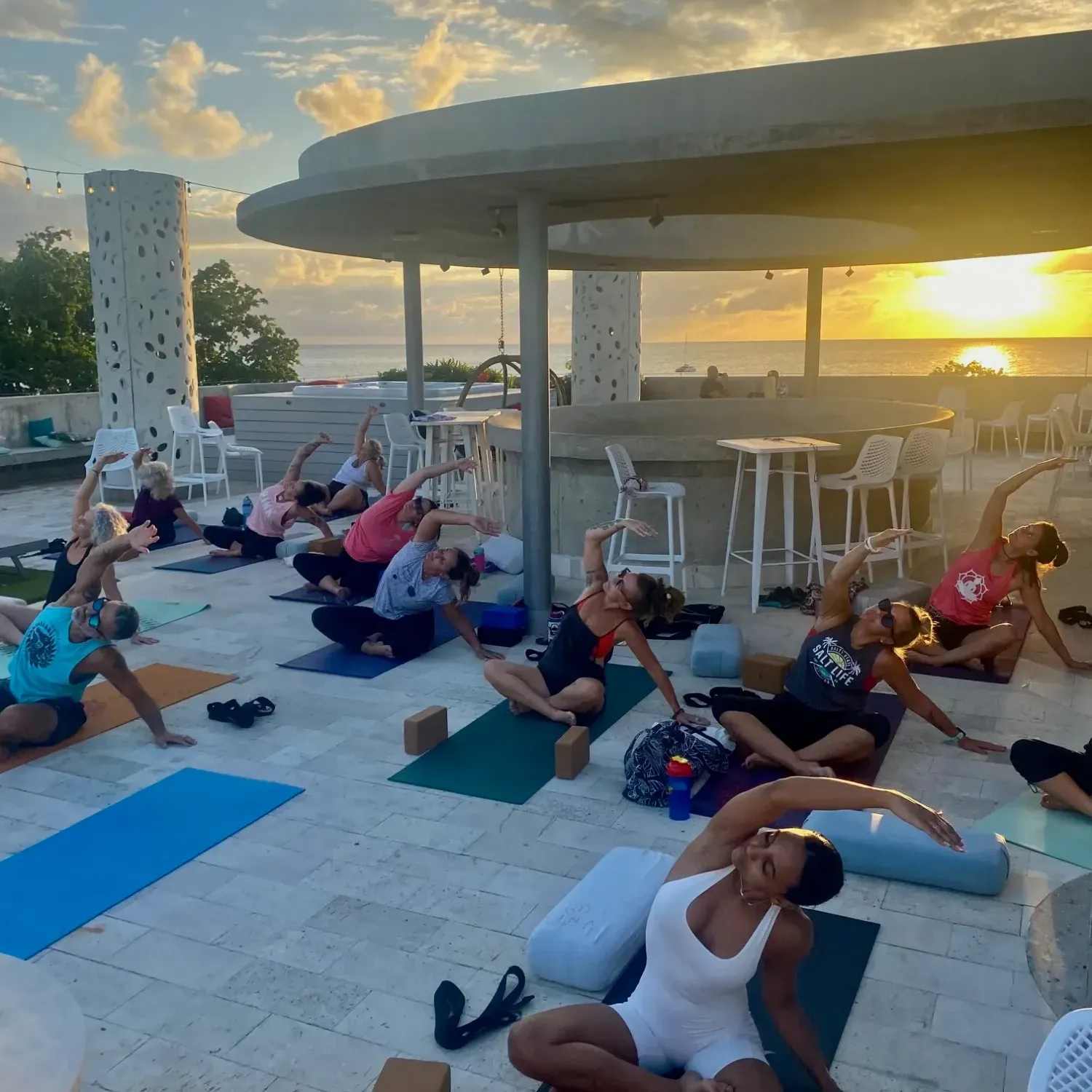 Group of people practicing yoga on mats on a rooftop terrace with ocean views during golden hour sunset.