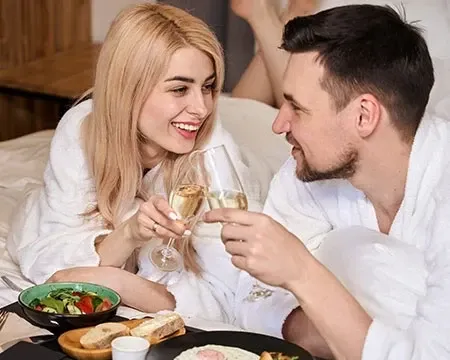 A couple in bed shares a romantic anniversary moment with champagne and food on tray.