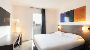 Clean hotel room with a bed, desk, chair, lamp, and TV at Appart’Hôtel Hévéa.