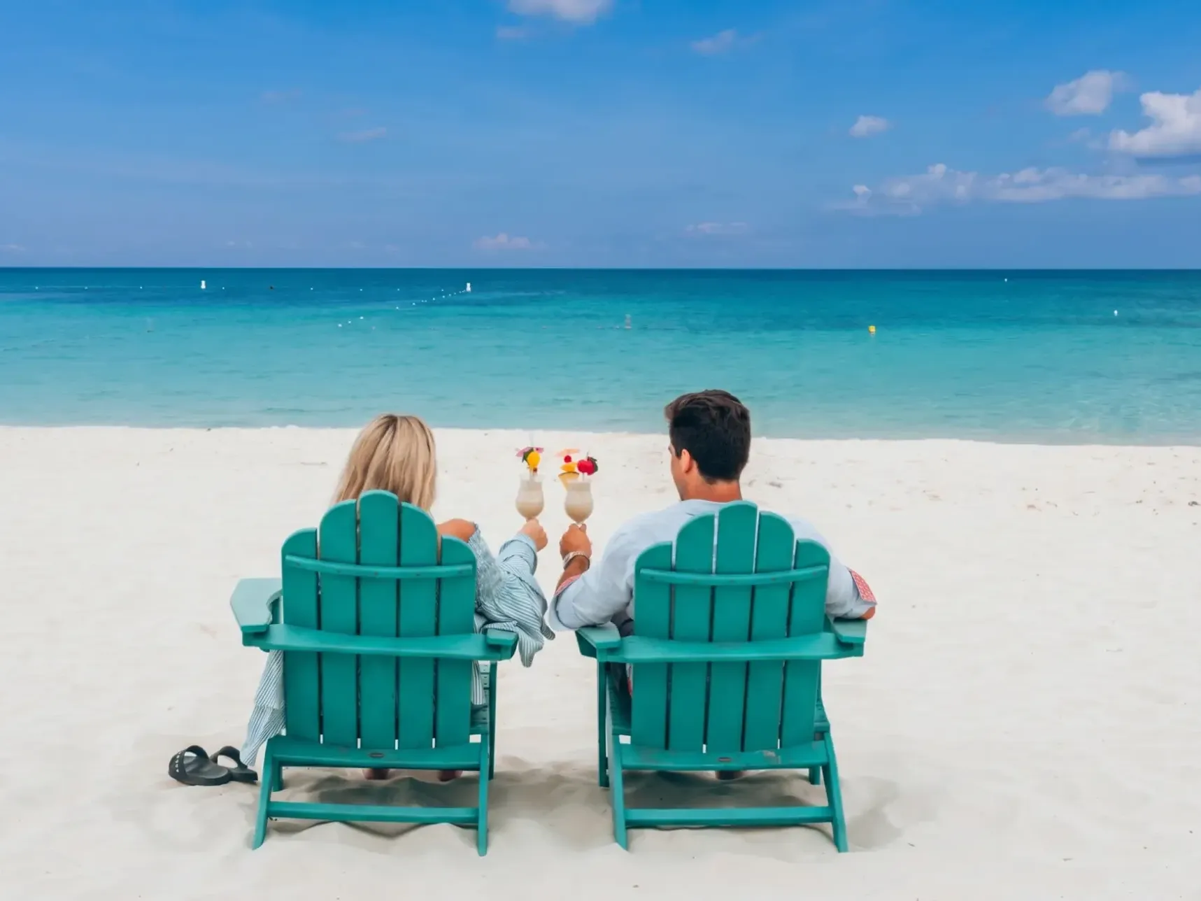 Couple relaxing in turquoise beach chairs with tropical drinks on white sand facing calm blue ocean waters