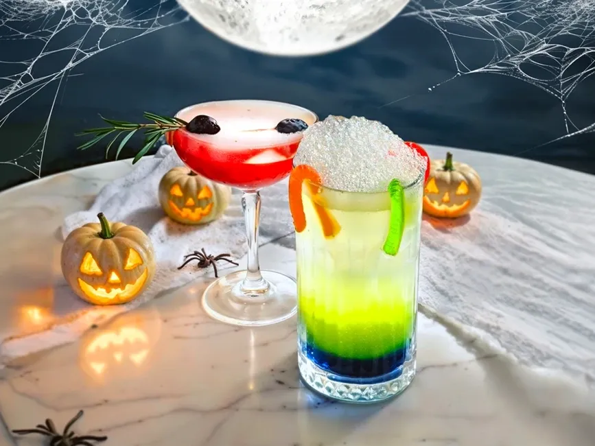 Halloween-themed cocktail and mocktail with spider webs, pumpkins, and full moon in the background
