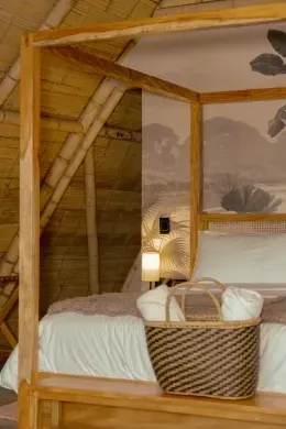 Four-poster wooden canopy bed with a woven basket, folded pillows, and lit lamp under a thatched roof at Unika Hotels
