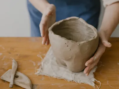 Person's hands holding a raw clay bowl on a wooden table at Good Times DIY Pottery & Tufting Studio.