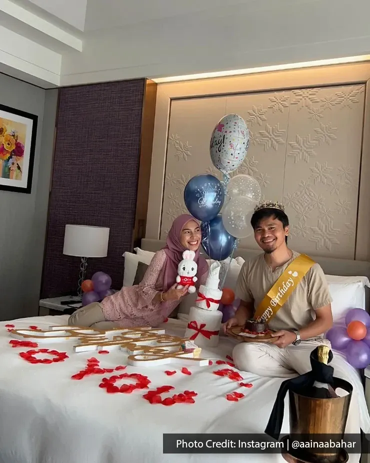 A couple enjoys a birthday celebration on a bed surrounded by colorful balloons and a delicious cake.
