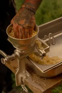 Tattooed hand pressing yellow corn kernels into a manual grinder over a stainless steel food pan outside at Unika Hotels