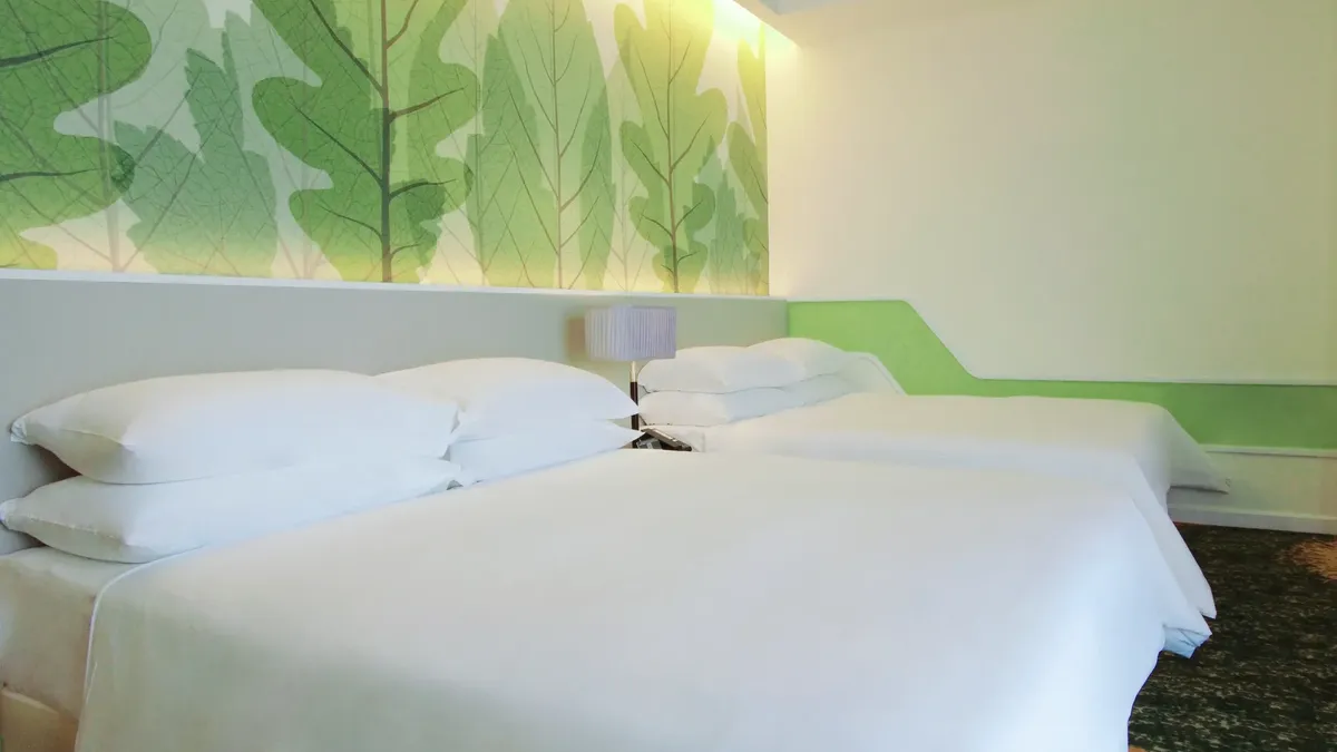Two white beds with pillows, green leafy wall decor, and a lamp in a hotel room.