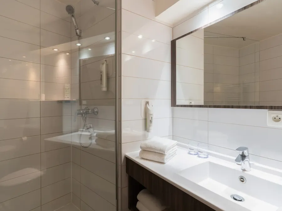 White tiled bathroom with a shower, sink, and mirror at Appart’Hôtel Hévéa, Valence.