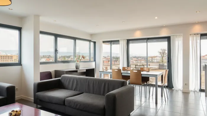 Spacious living area with couch and dining table overlooking city at Appart’Hôtel Hévéa.