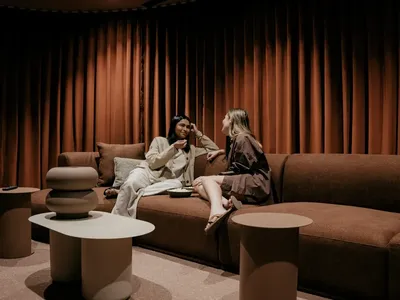 Two women chatting on a brown couch with brown curtains and tables at Caulfield Place.