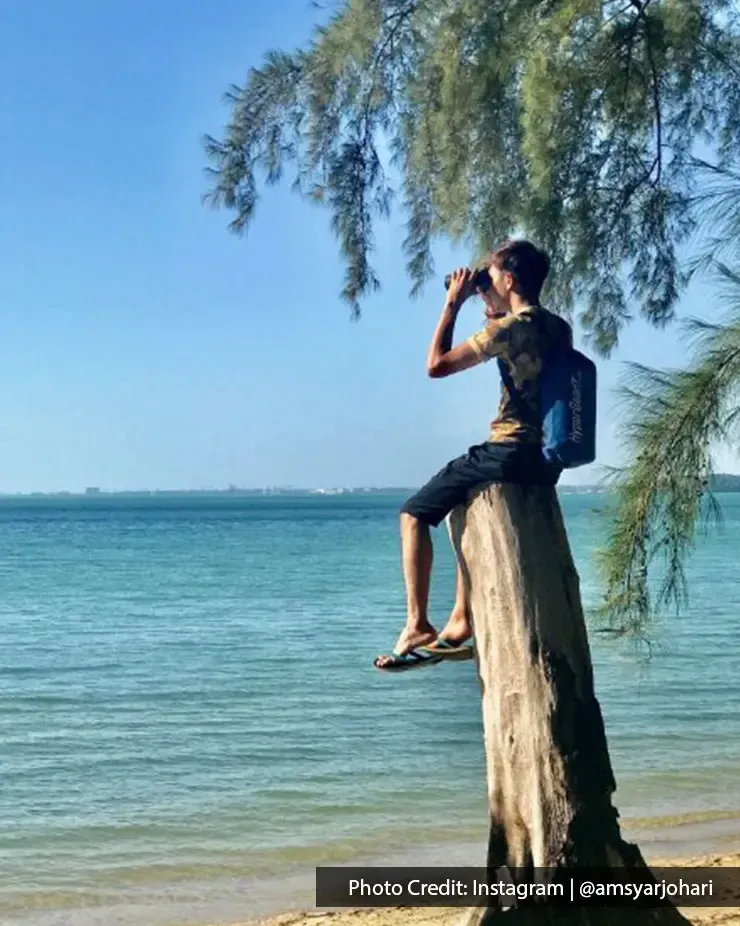 Man with backpack sitting on tree trunk at the beach overlooking the sea - Lexis Hibiscus Port Dickson