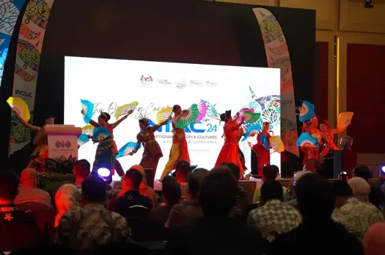 A diverse group of performers in vibrant costumes stands on stage at INTAC