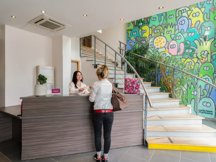 Woman checking in at Appart’Hôtel Hévéa front desk near colorful mural and staircase.