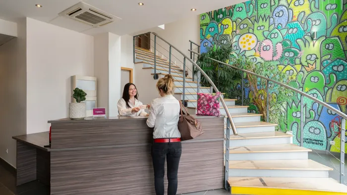 Woman checking in at Appart’Hôtel Hévéa front desk near colorful mural and staircase.